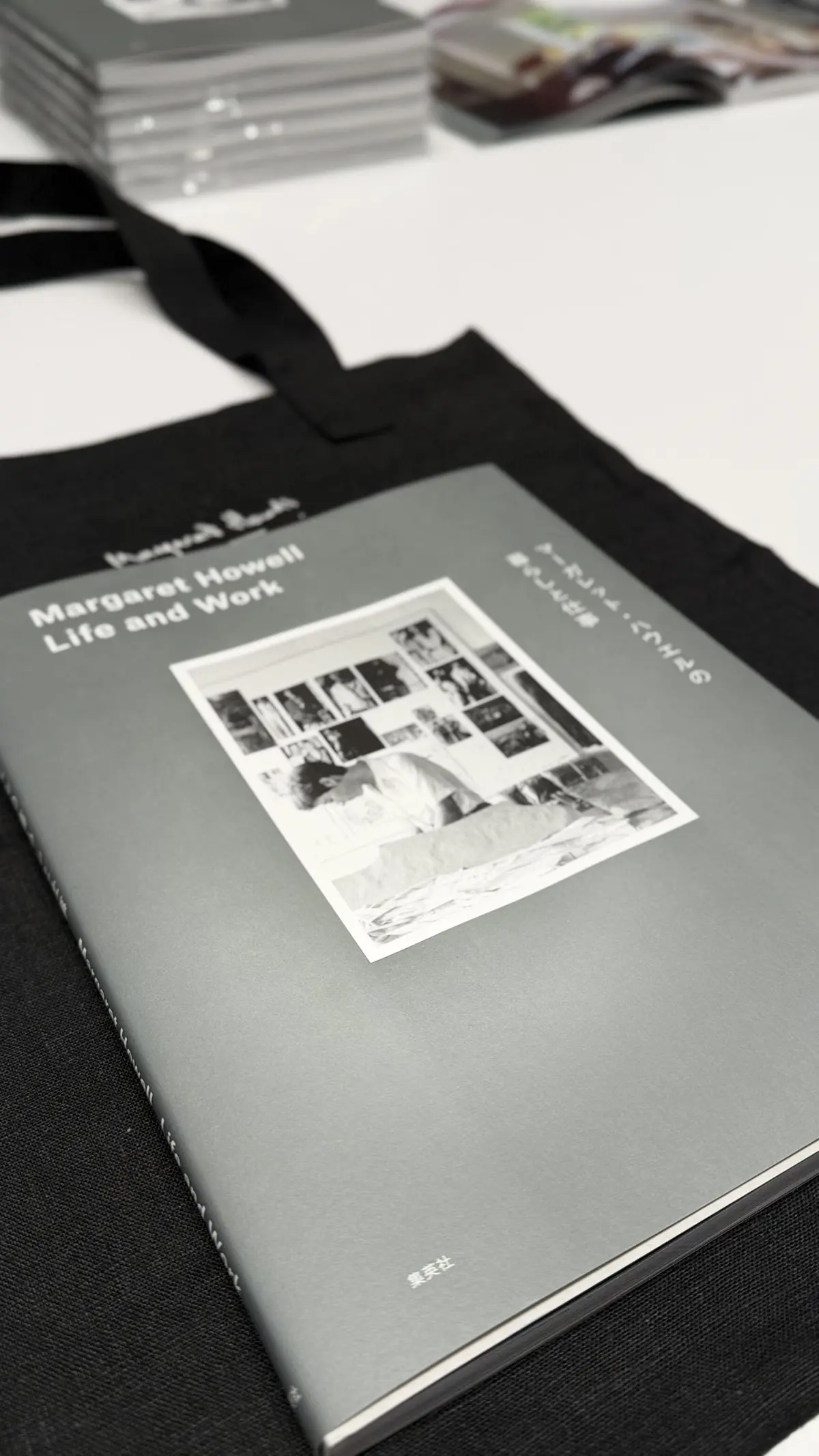 灰色の表紙に「Margaret Howell Life and Work」と白文字で表記された本。表紙にはモノクロ写真が数点コラージュされており、本が机の上に置かれている。背景には黒い布と、別の本が数冊積まれている。