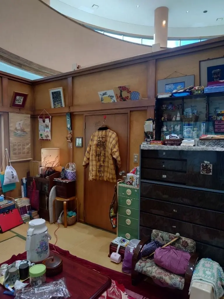 木目調の壁に装飾品が飾られた部屋。中央に黄色の着物姿のマネキン、右側にショーケース、手前にテーブルと座布団が置かれている。床は畳で、全体的に生活感のある空間。