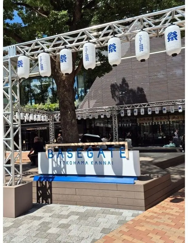 横浜関内の「BASEGATE」と書かれた看板。白い鉄骨のフレームに、白と青の提灯が飾られている。背景には、木々と近代的な建物が見える。