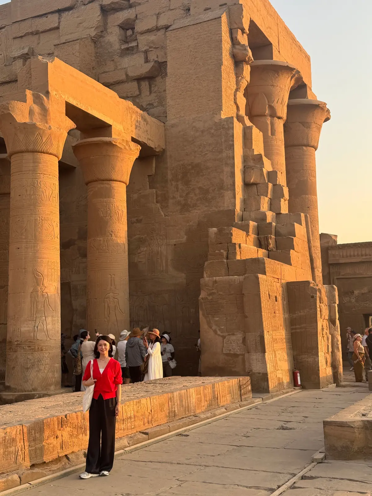 エジプトの古代遺跡、ルクソール神殿の柱の前で立つ女性。赤色のトップスに黒いパンツ姿。神殿はベージュ色で、多くの柱と彫刻が施されている。夕焼けの光が神殿を照らしている。