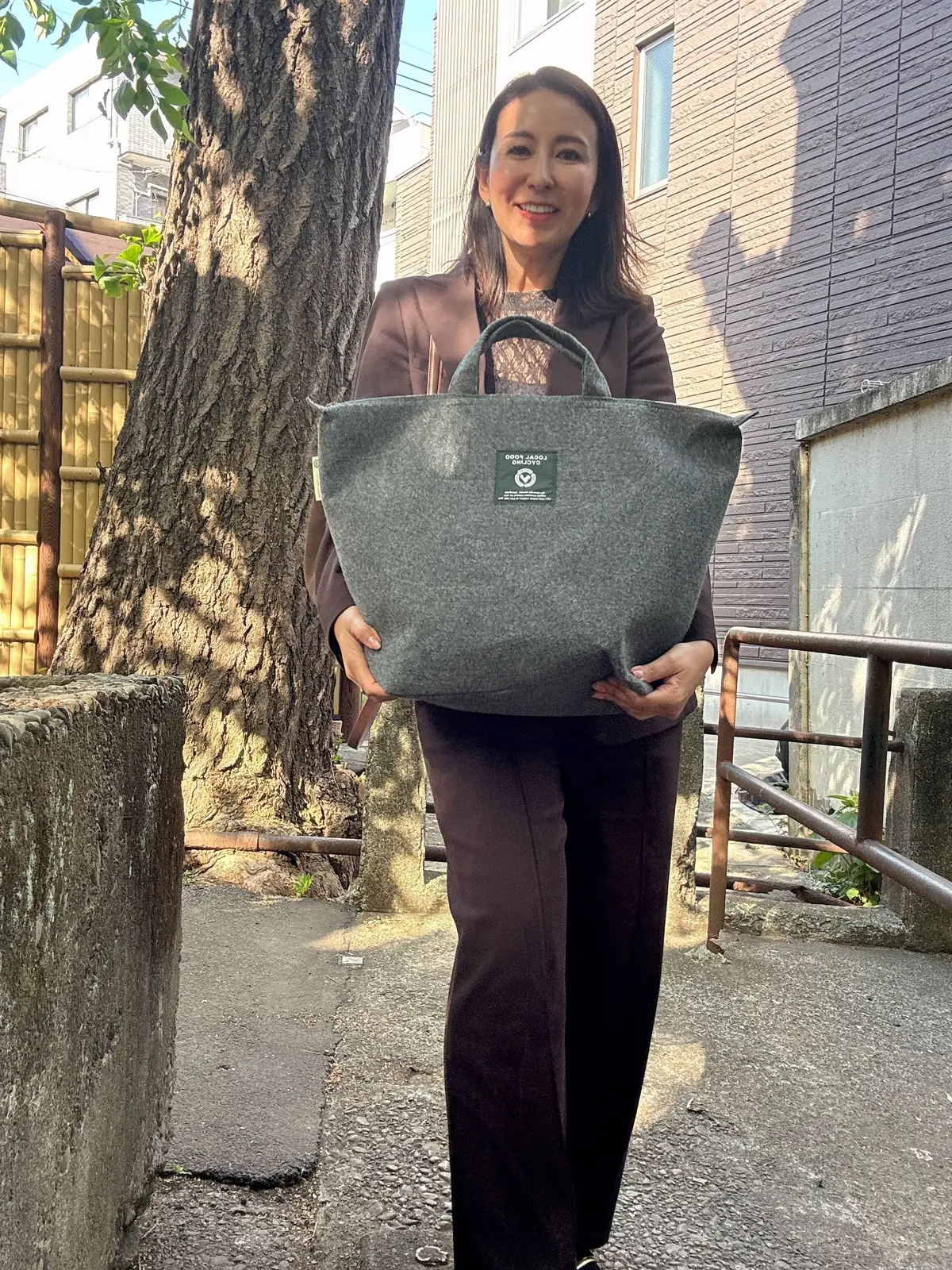 女性が大きなグレー色のトートバッグを手に持つ。背景には木や建物が見える。