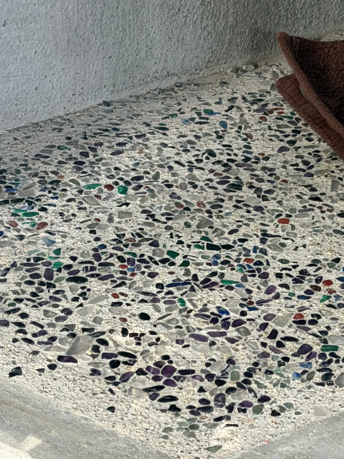 白いセメントの表面に、様々な色（青、緑、紫、赤など）の小片が散りばめられたテラゾー加工の床。右端には茶色の布が見える。
