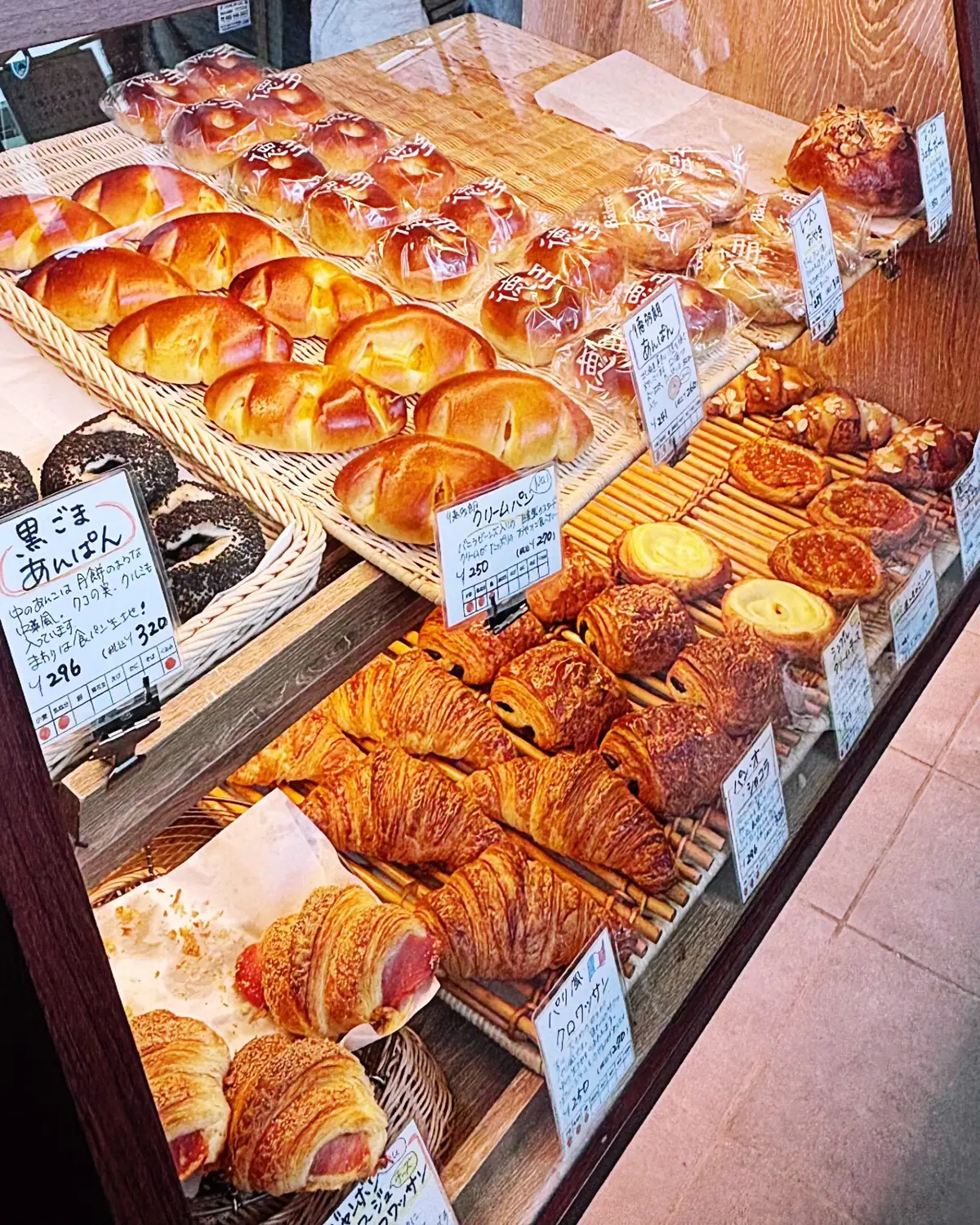 パンと菓子類が並ぶ店内のショーケース。クロワッサン、ドーナツ、パンが木製トレイに整列し、価格表が添えられている。