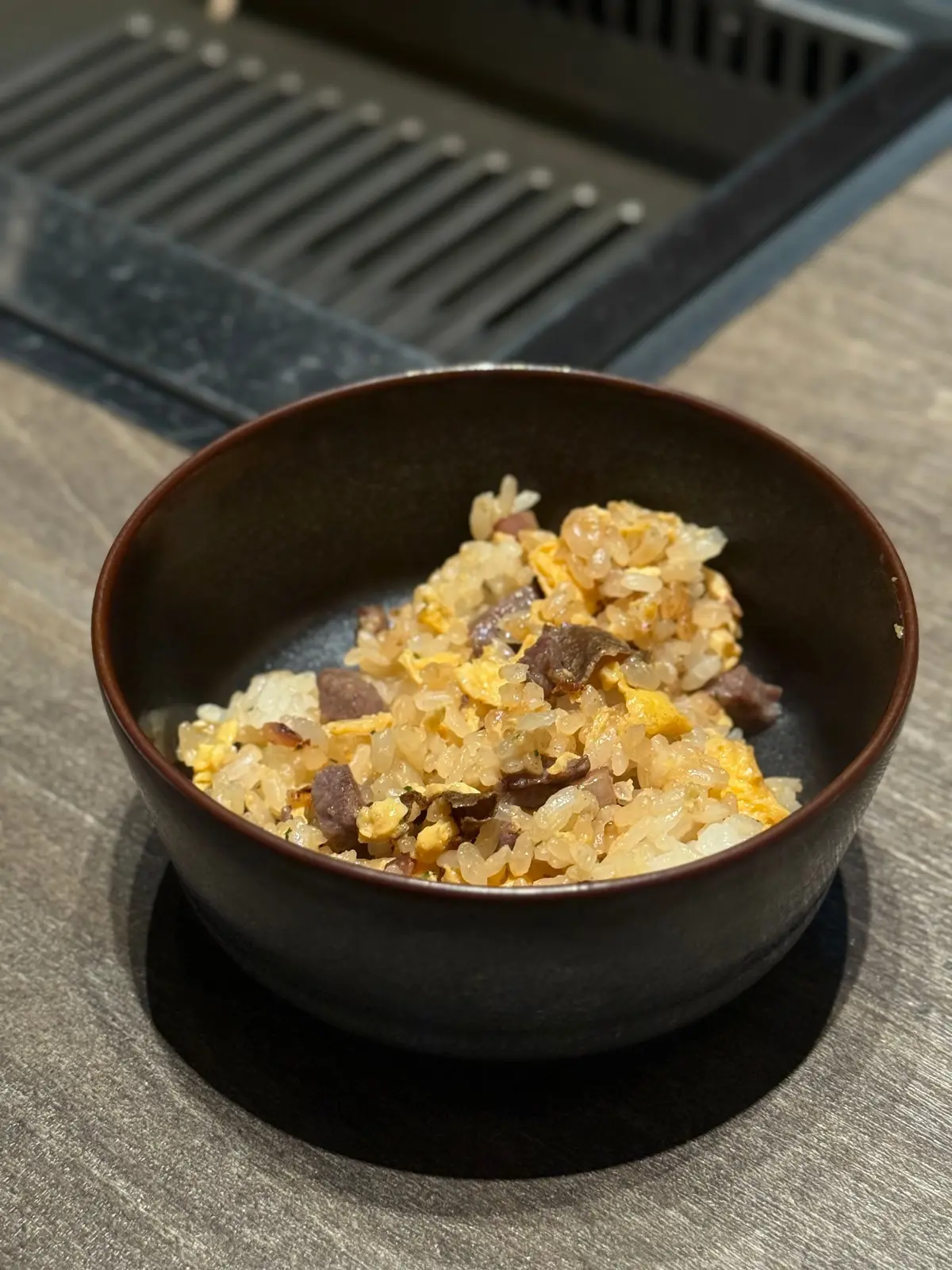 茶色の陶器のボウルに入った、卵とご飯を混ぜた炒飯。黒っぽい具材が散らばっている。背景には鉄製のグリルが見える。
