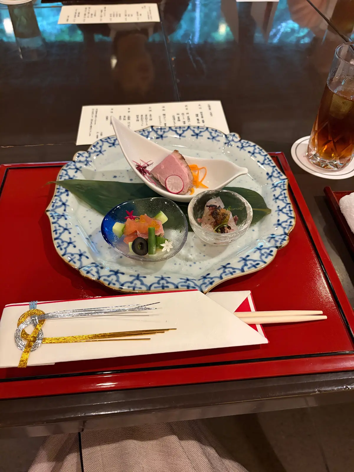 青い皿に盛り付けられた、3種類の日本料理。左はピンク色の肉、中央はフルーツとオリーブ、右は魚介の品。白磁のソース入れと、黄色の箸、赤い箸置きが添えられている。