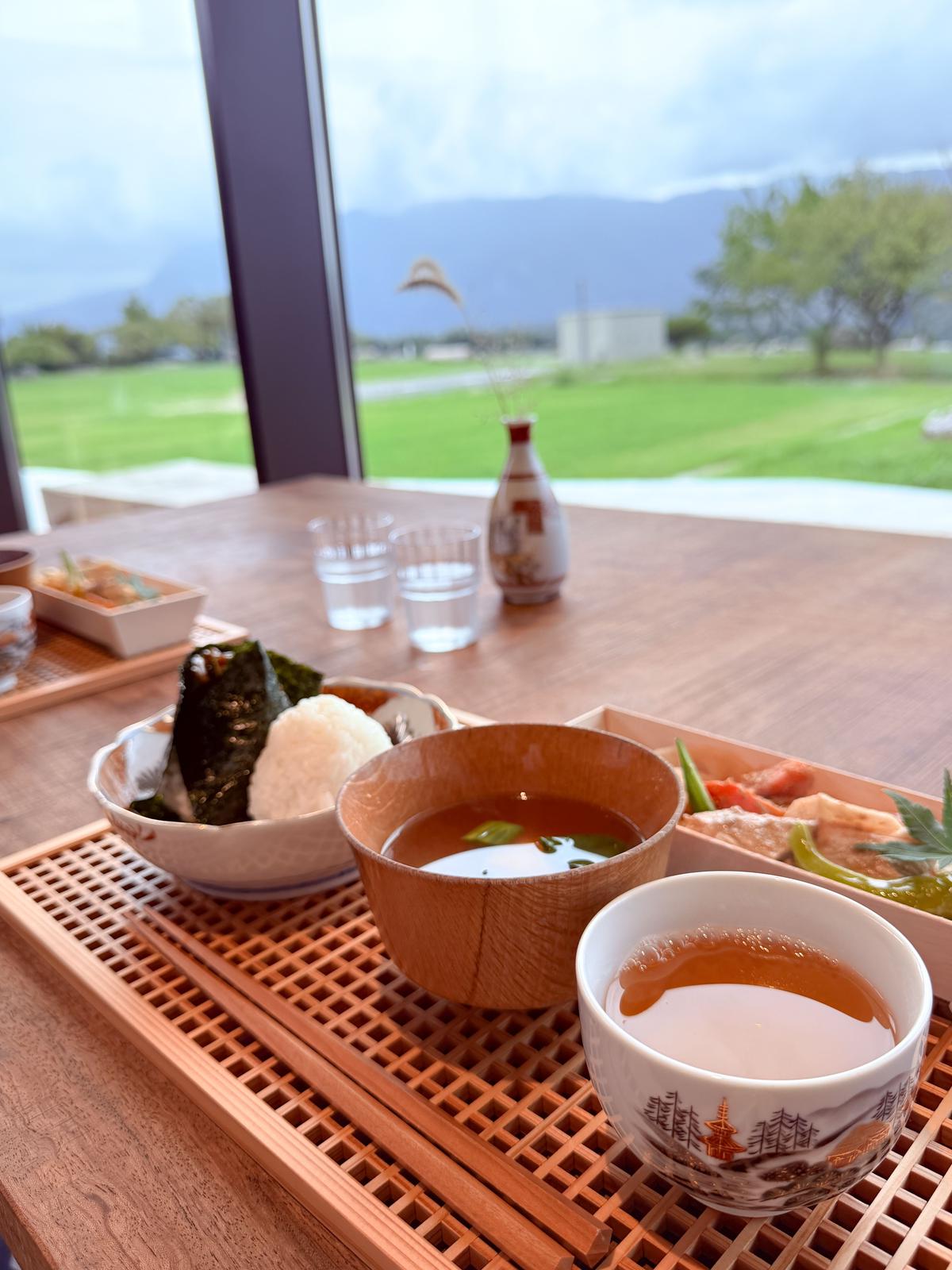 木製のトレイに盛り付けられた和食。海苔に巻かれたおにぎり、お味噌汁、お漬物、茶碗に入ったお茶。背景には緑の芝生と山々が見える窓。