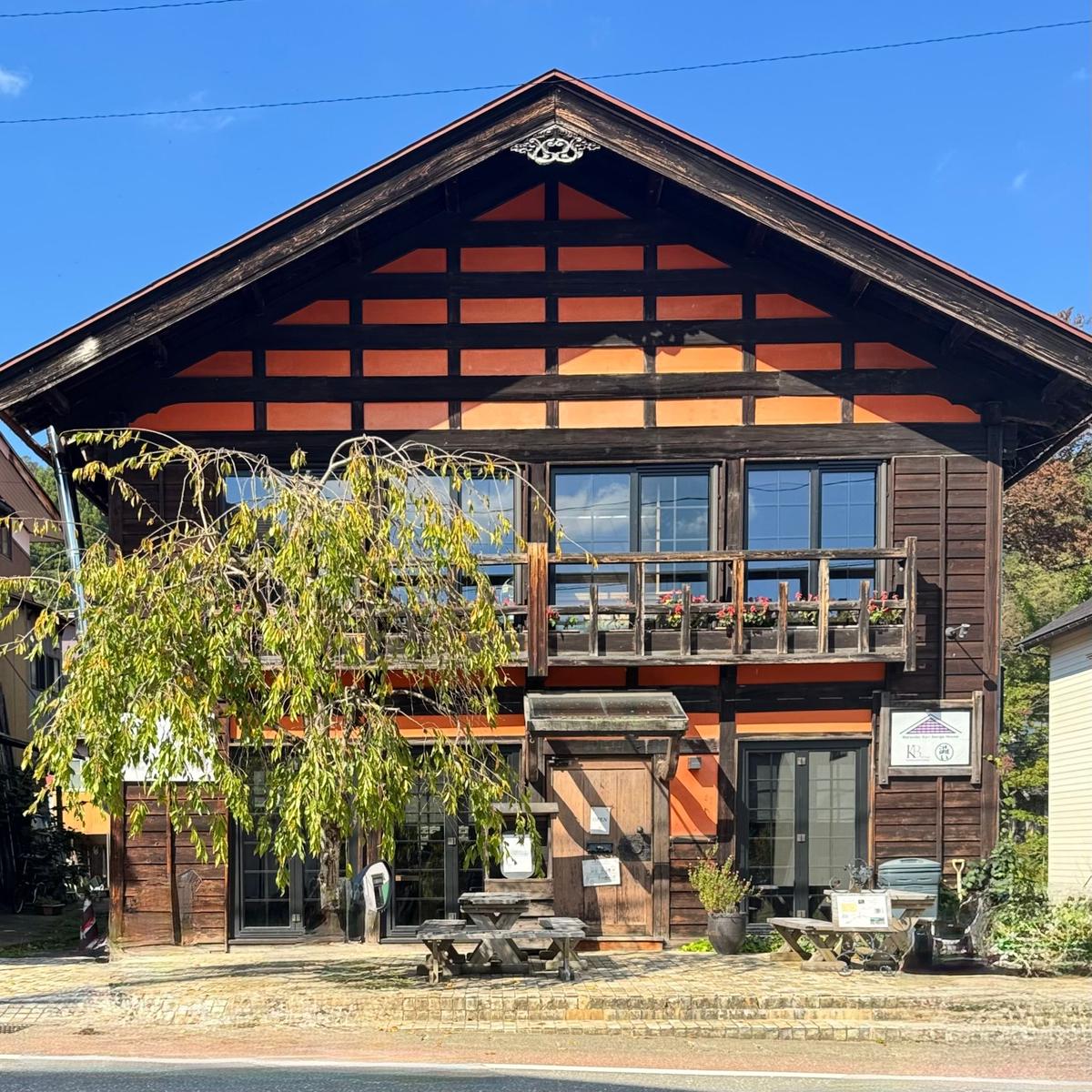 木造の2階建て建築。屋根は濃い茶色で、梁がオレンジ色に塗られている。窓辺には花が飾られ、木製のバルコニーがある。正面には木製の扉と看板が設置されている。