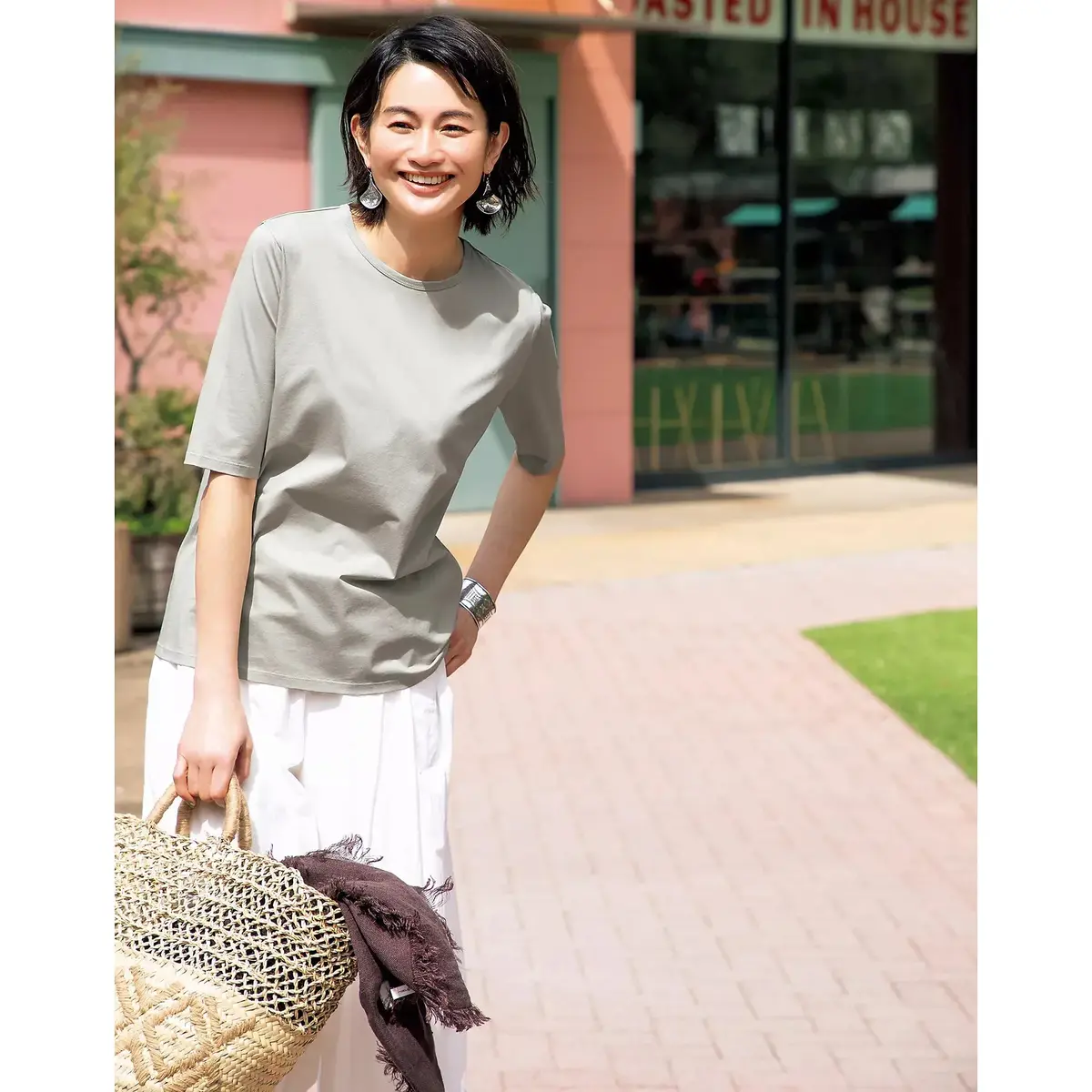 女性が白い半袖トップスと青いロングスカートを着て、屋外の緑豊かな庭園で微笑んでいる。手には淡い青いショルダーバッグを持っている。背景には木や低木が広がっている。