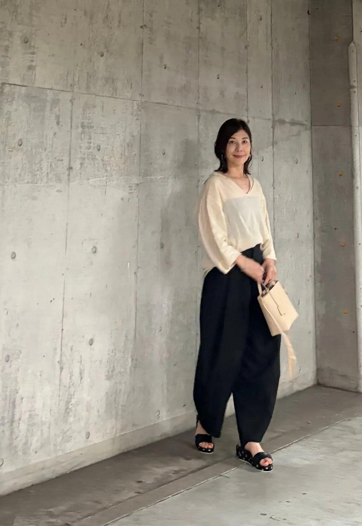 コンクリート壁に寄り添って立つ女性で、ベージュの長袖シャツと黒いワイドパンツを着用し、ベージュのトートバッグを右手に持ち、黒いサンダルを履いている