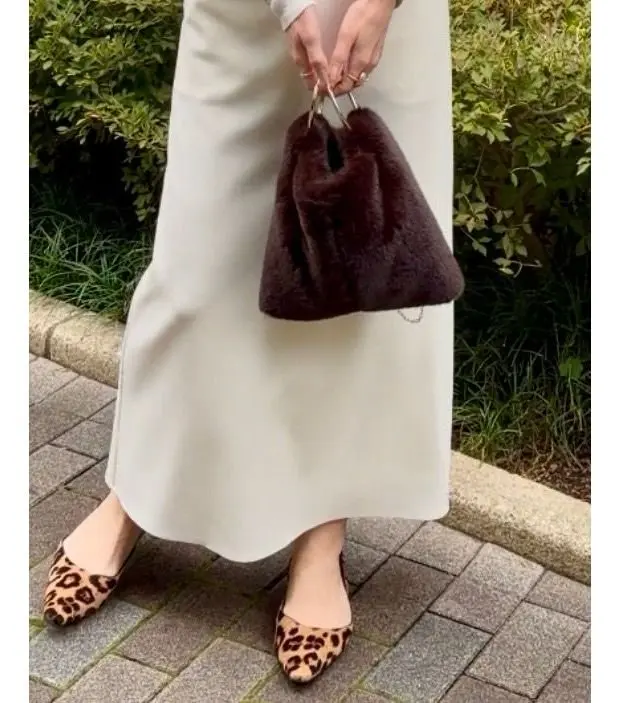 オフホワイトのロングスカートと、レオパード柄のフラットシューズを履いた女性。手にはダークブラウンのふわふわしたミニバッグを持っている。背景には緑の植物が見える。