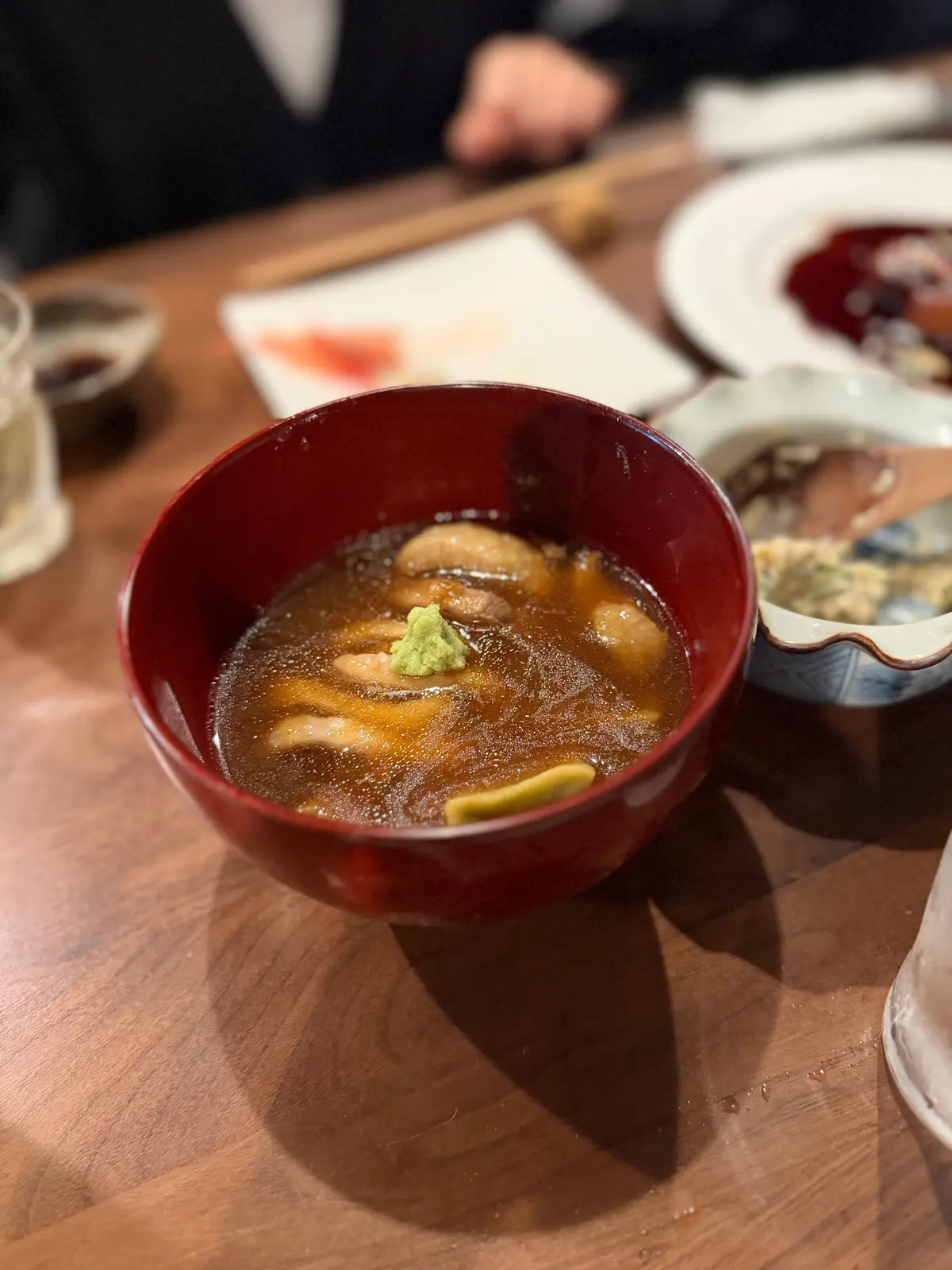 赤い漆塗りの椀に入った、薄い茶色の汁物。中央には緑色の薬味（わさびか？）が置かれ、椀の縁には金色の粉が散らされている。木製のテーブルに置かれ、背景には食器や紙ナプキンが見える。