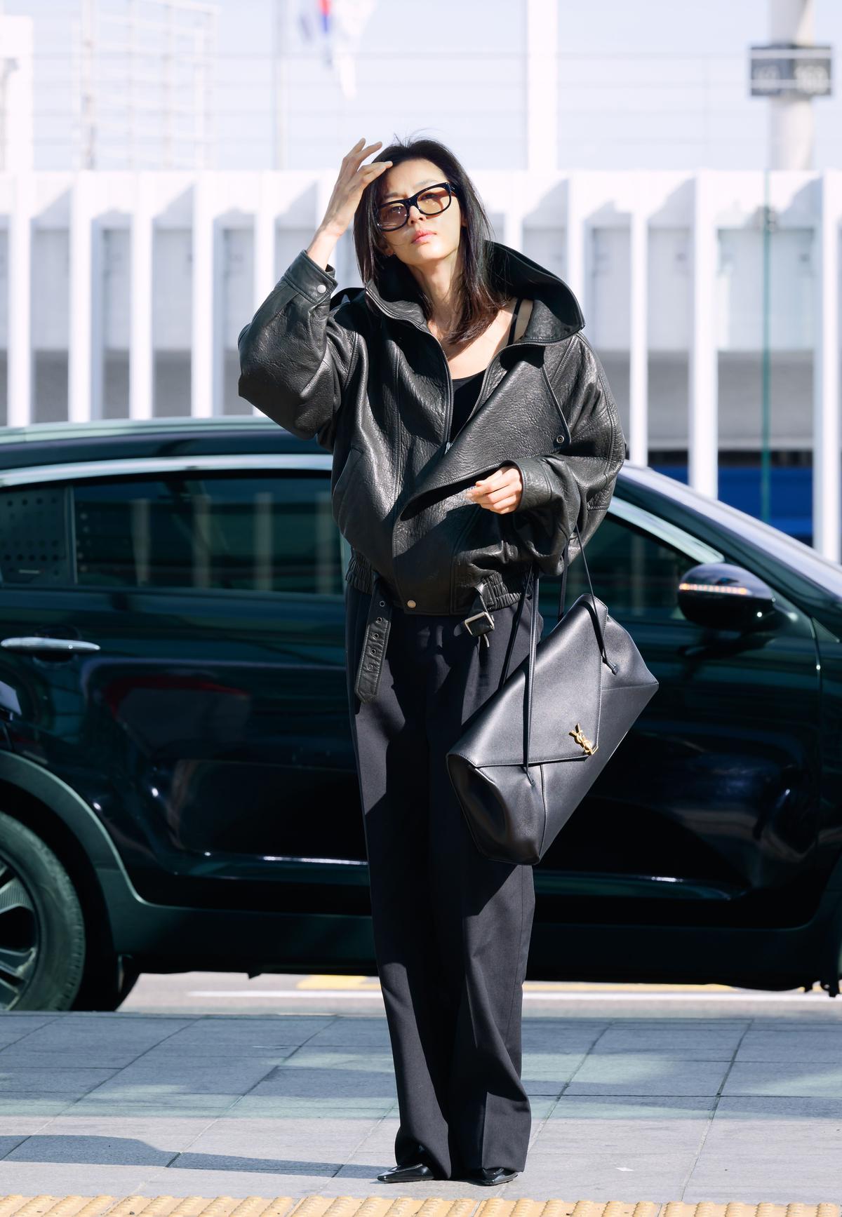 黒いレザージャケット、黒いパンツ、黒縁メガネを着用した女性が立っている。黒いハンドバッグを肩にかけ、背景には黒い車と白い建物がある。