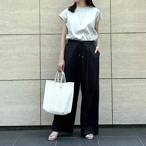 白いネックレスをつけた女性が、黒いドローストリングパンツと白いシルクのトップスを着て立っている全身写真。右手に白いトートバッグを持ち、背景はベージュのタイル壁。