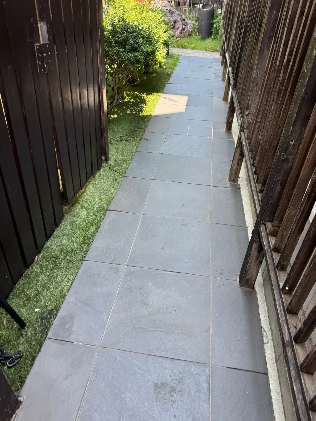 左右に木製の柵が並ぶ、幅の狭い通路。通路は灰色の石畳で、奥には草むらと緑の植物が見える。