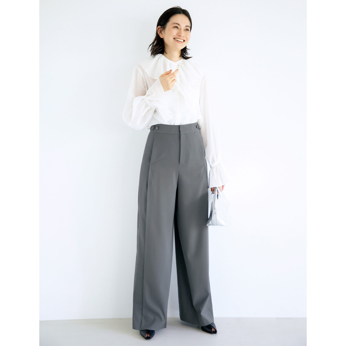 白いフリルシャツにグレーのワイドパンツを合わせた女性。笑顔でこちらを見ている。右手にシルバーのバッグを持つ。背景は白。