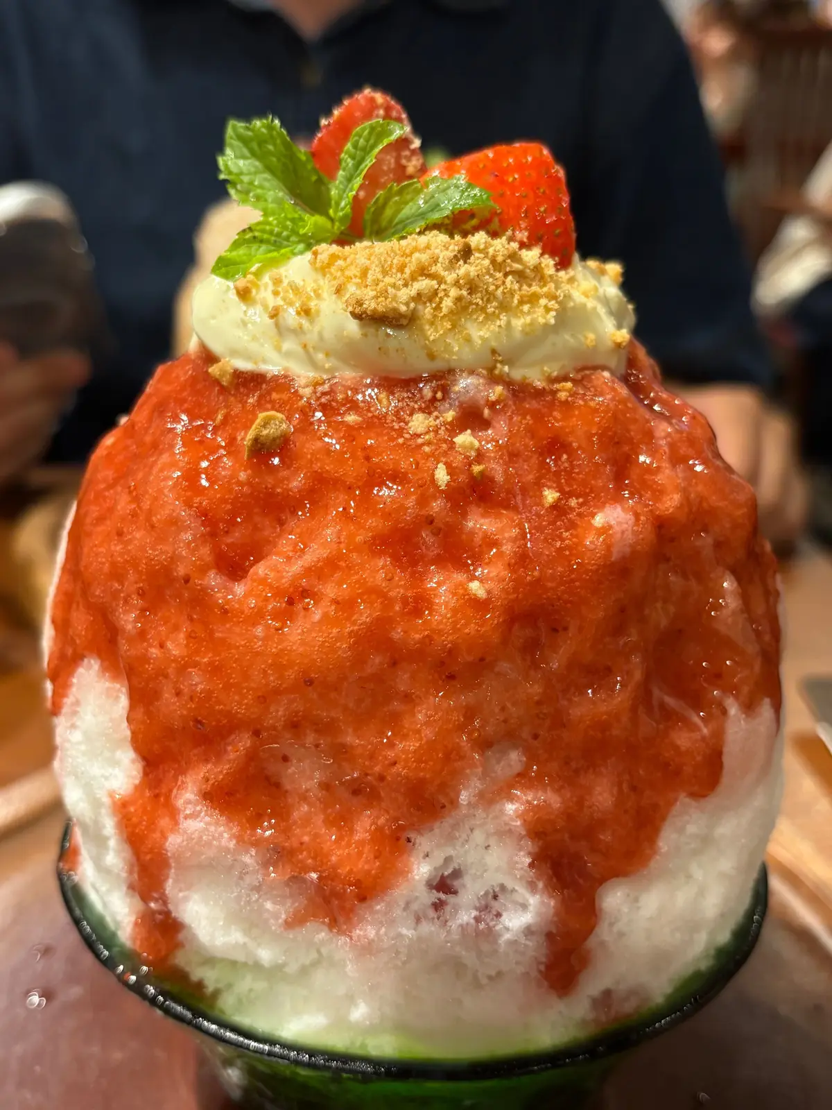 緑色の器に入った、高さのあるかき氷。赤色のイチゴソースと、砕いたナッツがたっぷり。中央にはクリームとイチゴ、ミントの葉が飾られている。