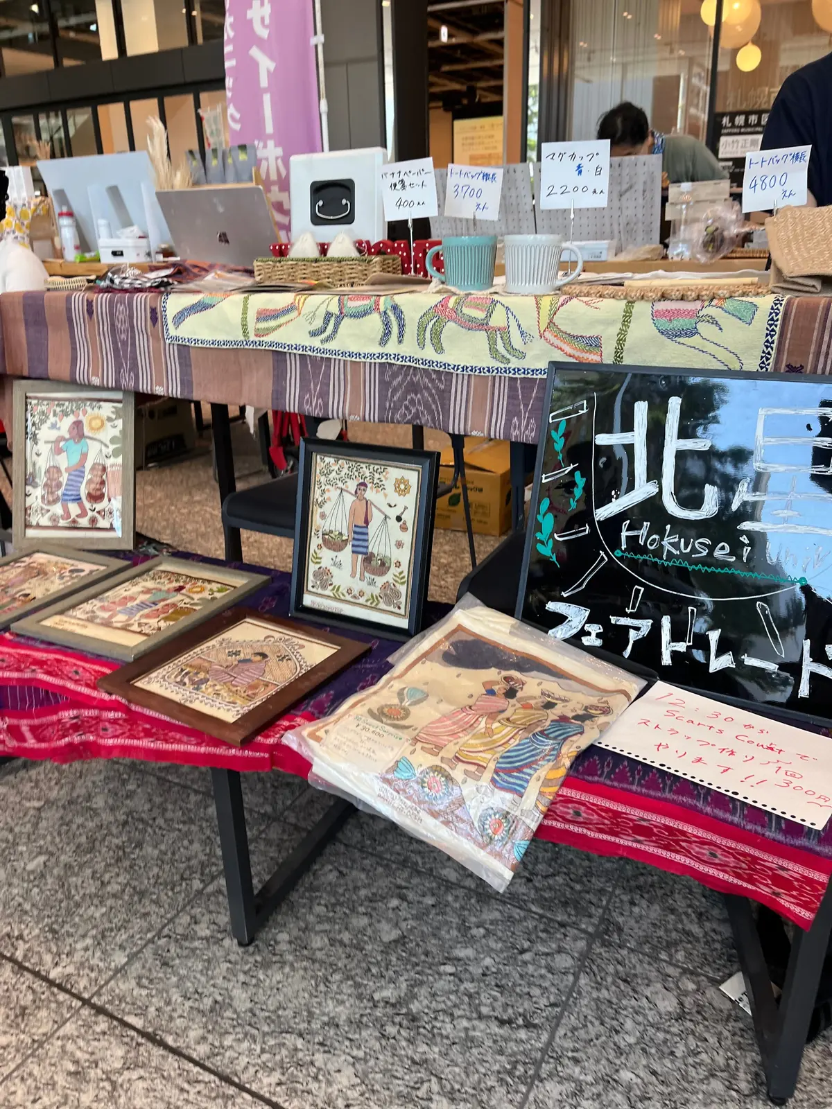 多様な絵画やイラストが並ぶアートマーケットのブース。色鮮やかな布で覆われたテーブルには、絵画やイラスト、価格表示の看板、飲み物カップが置かれている。手描きの絵画やポスターが展示され、観客が作品を眺めている。