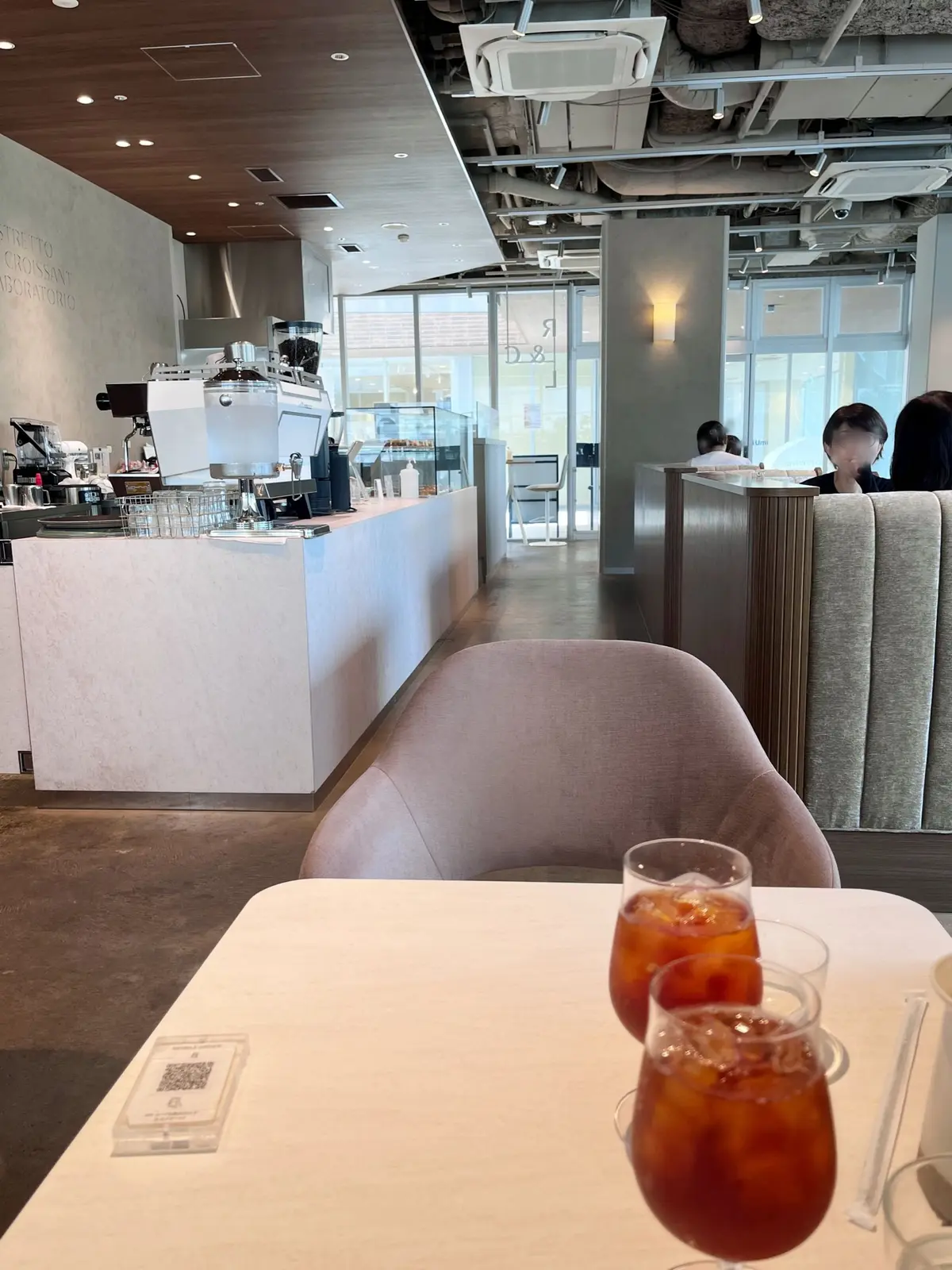 カフェ「リストレット＆クロワッサン研究所」のテラス席。丸い大理石のテーブルと、黒い革張りの椅子が2脚置かれている。大きなガラス窓の向こうには緑豊かな植栽が見える。