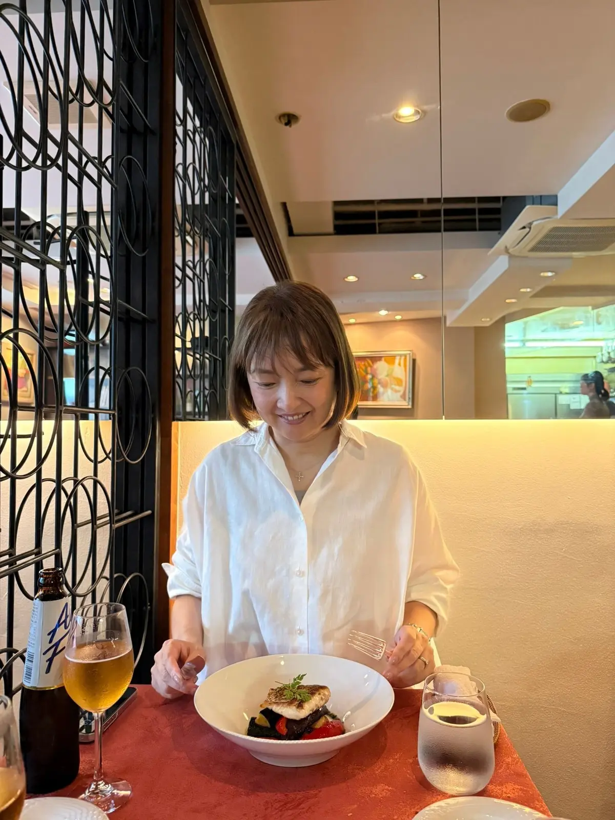 レストランのテーブル席に座り、白いシャツを着た女性が魚料理を食べている。背景には装飾的な黒い仕切りと、厨房の様子が見える。