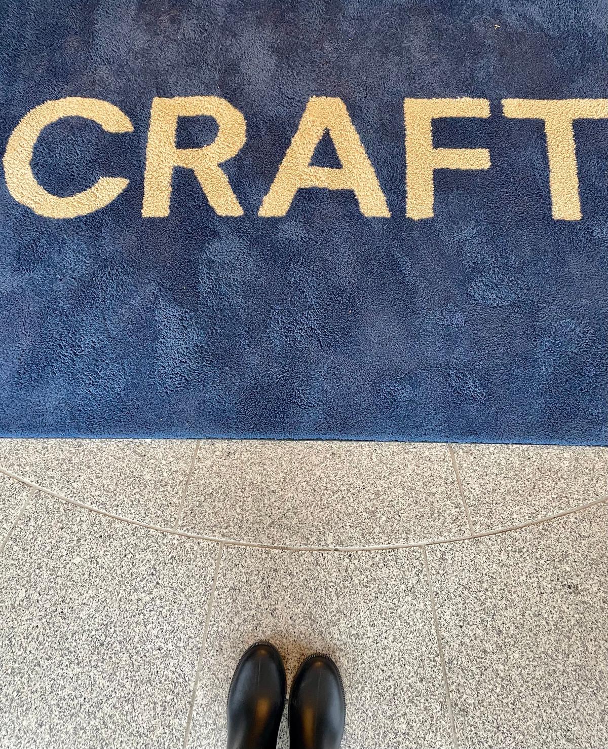 青い敷物の中央に「CRAFT」の文字が大きく刺繍されている。敷物の下にはベージュ色のタイルが敷かれており、足元には黒いブーツが見える。