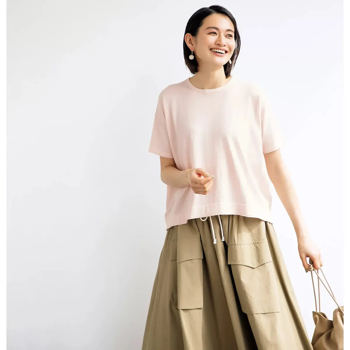 ピンクの半袖Tシャツとベージュのワイドパンツを着た女性が、笑顔で白い背景に立っています。右手にはベージュのショルダーバッグを所持しています。
