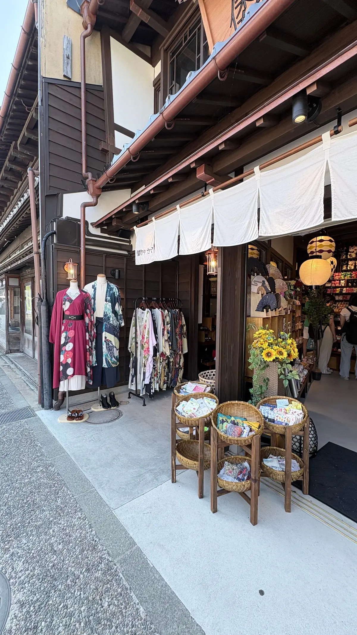 木造の建物沿いに並ぶお店の様子。白い布で覆われた出店には、着物や小物が展示され、藁の椅子に手拭いが置かれている。向こう側には、提灯や商品が並ぶ様子が見える。