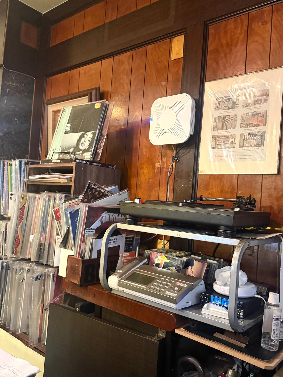 木製の壁に沿って設置されたレコード店のレジカウンター。カウンターにはレジ、電話、レコードが置かれ、右壁には複数のレコードジャケットが飾られている。左壁には黒板と、いくつかのレコードが立てかけられている。