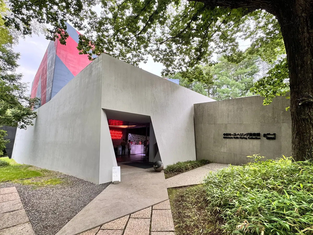 コンクリート打ち放しの現代美術館。赤いアクセントカラーの屋根と、館内展示の様子が映る入り口。緑豊かな木々が背景に茂る。