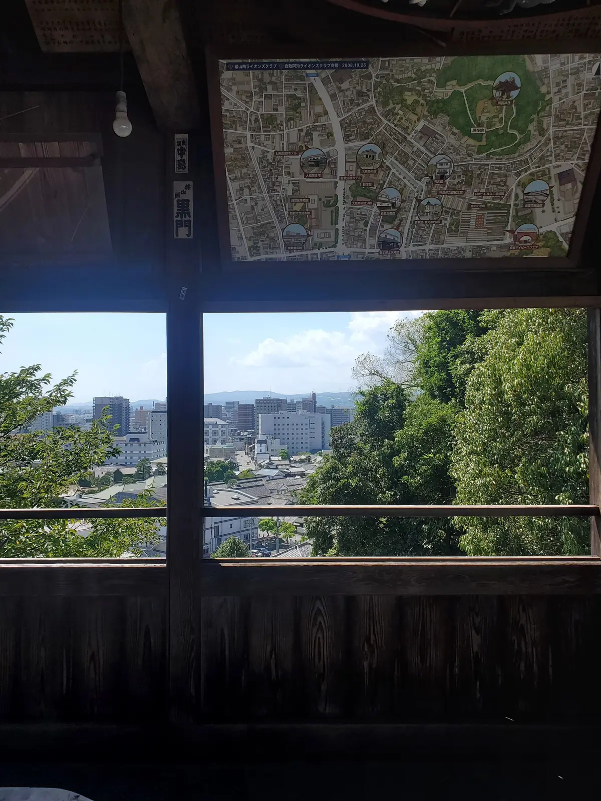 木製の観測所の窓から見える、緑豊かな丘と都市の風景。窓枠の上の壁には地図が貼られ、窓からは高層ビル群が望むことができる。
