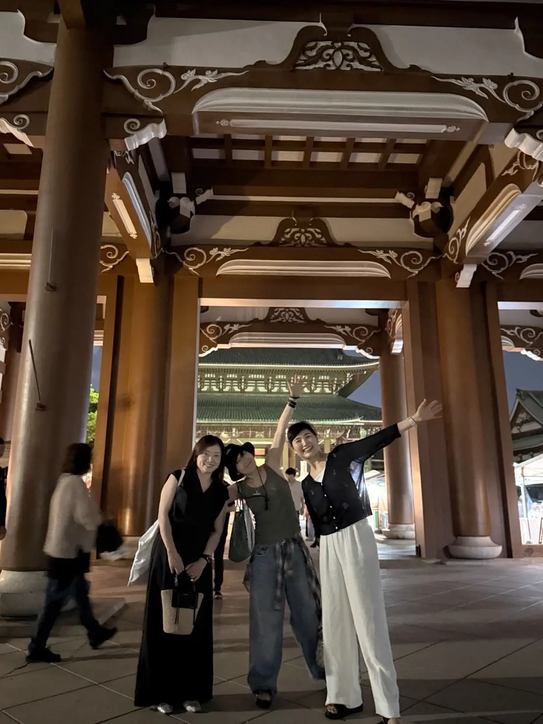 夜の寺院のアーチの下で、3人の女性が写っている。左から黒いワンピース、グレーのTシャツにジーンズ、白いパンツと黒いトップスを着ている。右側の女性が手を振っている。背景には、緑色の屋根の建物が見える。