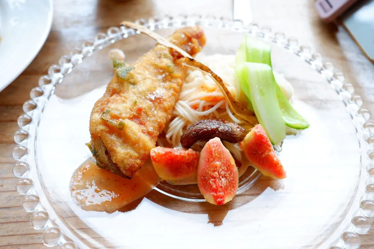 ガラス皿に盛り付けられた、揚げ魚と麺、フルーツの料理。魚は淡いピンク色で、麺は白。赤や緑のフルーツが彩りを添えている。