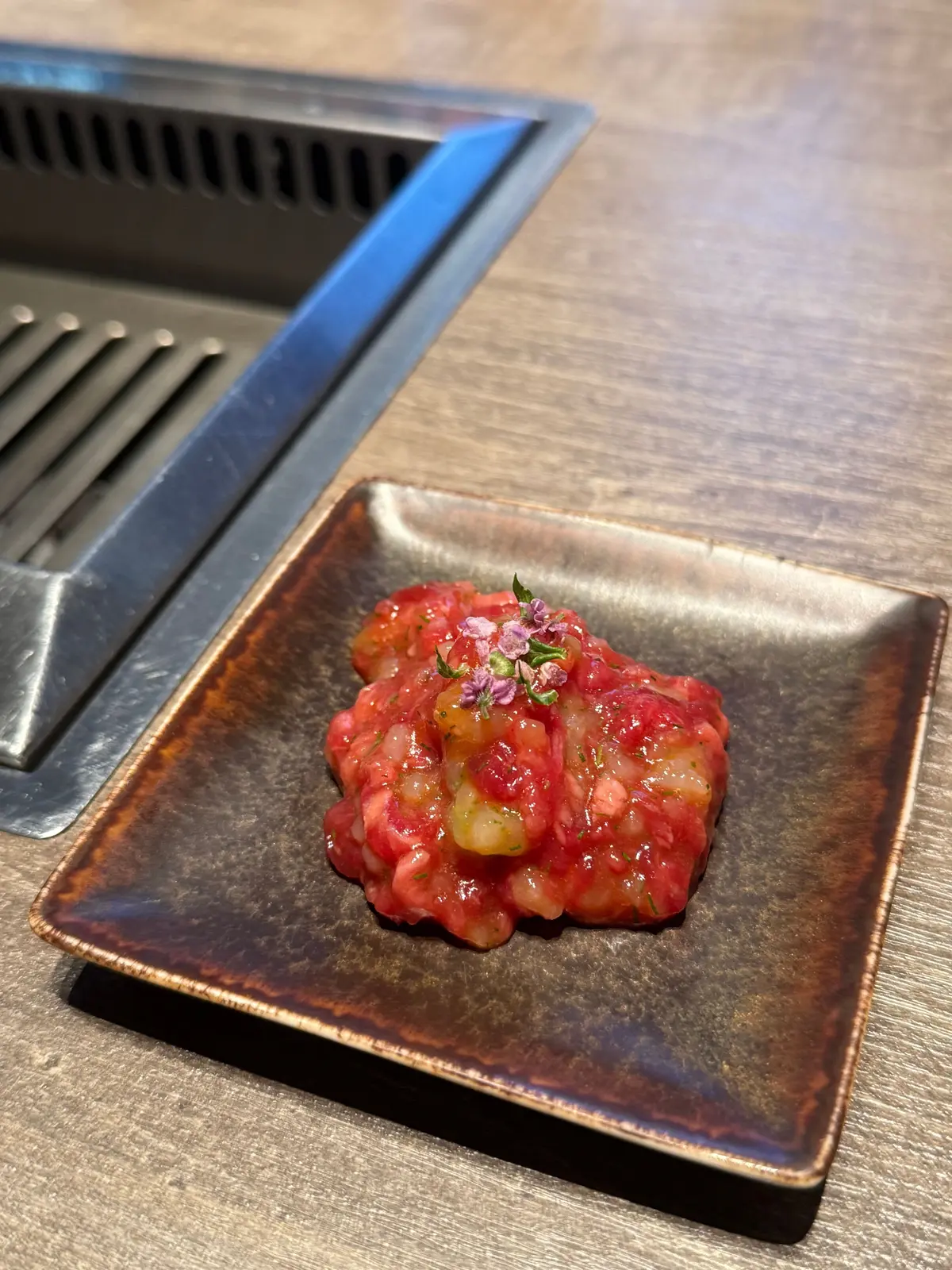 茶色の四角い皿に盛り付けられた、赤と緑色の細かく刻んだ料理。表面には小さな紫色の花が散らされている。皿の横には金属製のグリルが置かれている。