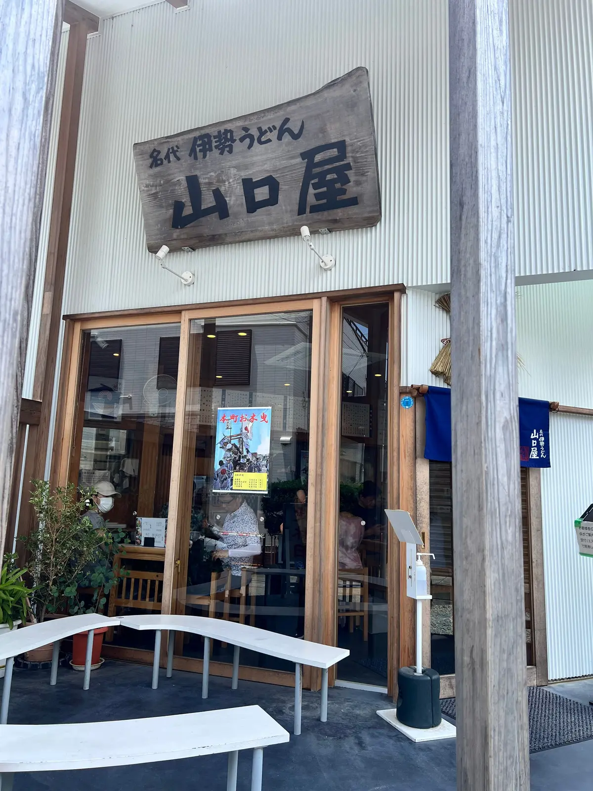 木製の看板が掲げられた、白い壁と金属製の外装の店舗。ガラス張りの入り口にはメニューらしきポスターが貼られ、テーブルと椅子が並べられている。