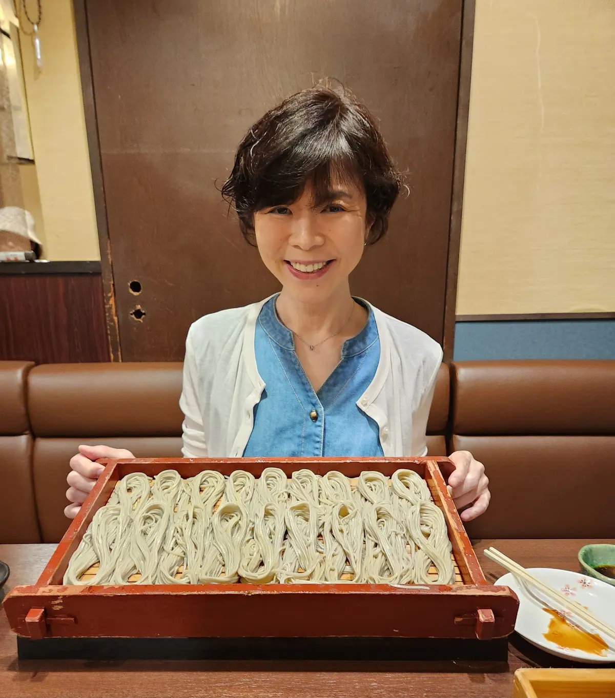 女性が手板に並べられた白いうどんを両手で持ち、笑顔でカメラに向かって見つめている。背景には木製の壁と茶色の革の座席が確認できる。