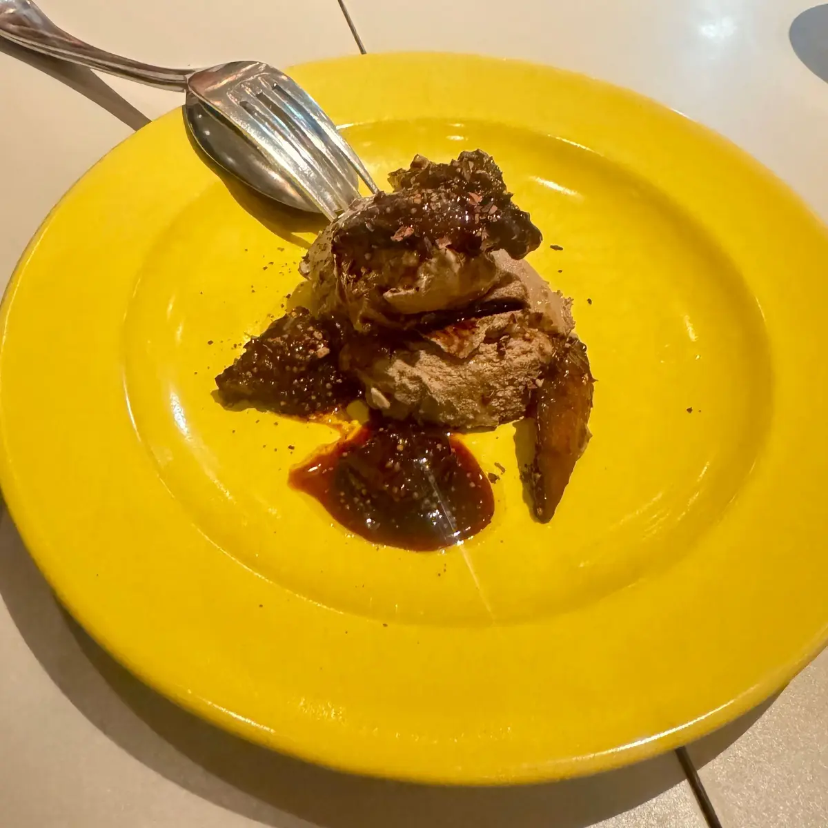 黄色い皿に盛り付けられた、チョコレート系のデザート。アイスクリームのような塊と、ソースをかけた果肉が添えられている。左にはフォークが置かれている。