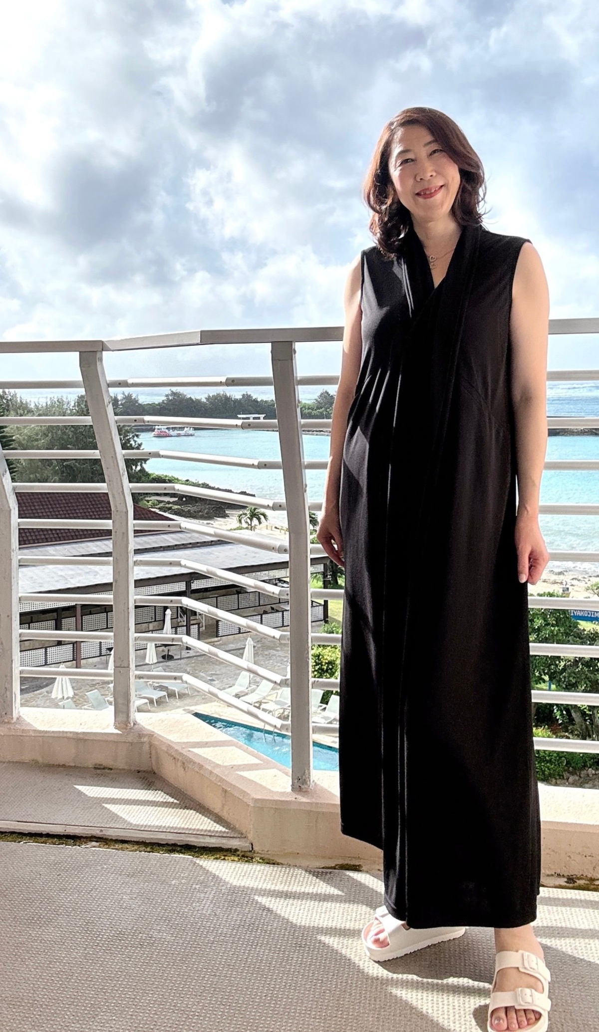 黒いワンピースを着た女性が、海を見下ろすテラスに立っている。背景には青い海と空、白い波打ち際、ホテルが見える。