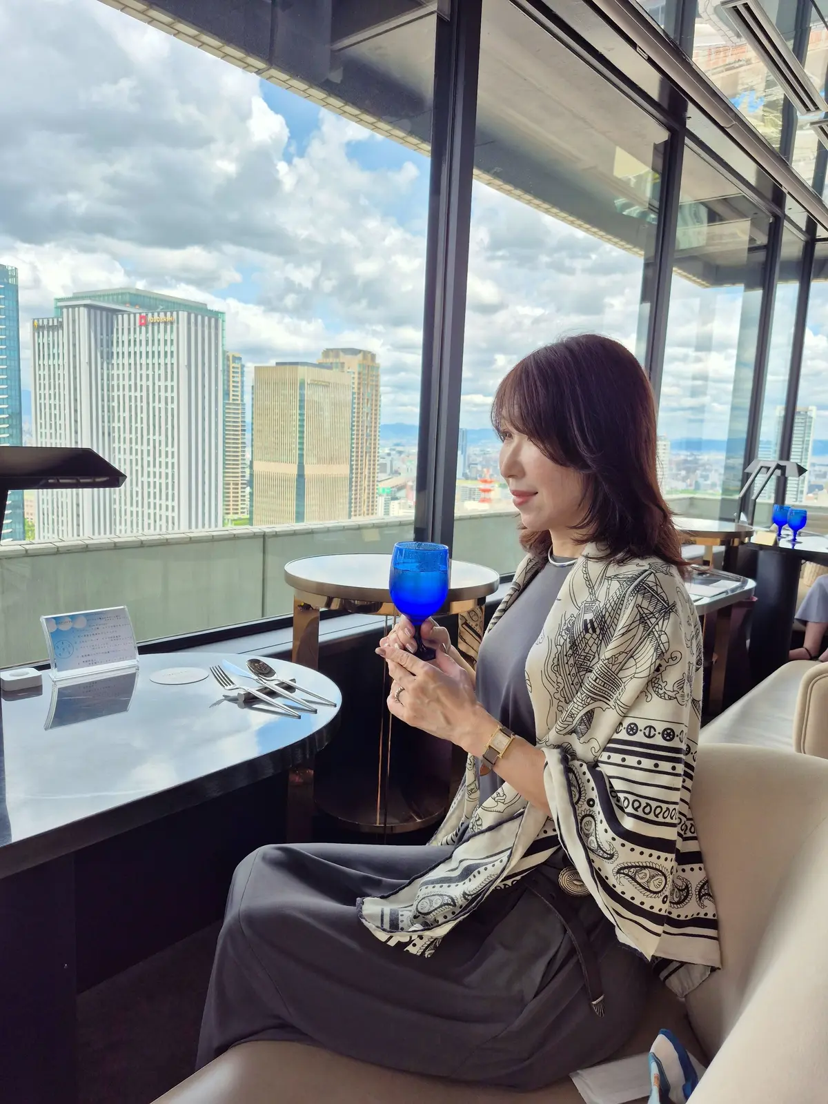 高層階のレストランで、女性が青いグラスを手に窓の外を見ている。背景には高層ビル群と雲のある空が広がる。女性はグレーの服に黒と白の柄のショールを羽織っている。