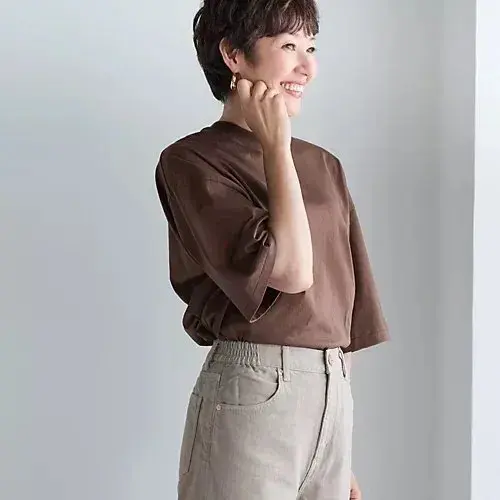 茶色のボックスTシャツに、ベージュのパンツを着用した女性が微笑んでいる。右手を首に当て、横を向いている。背景は白っぽい壁。