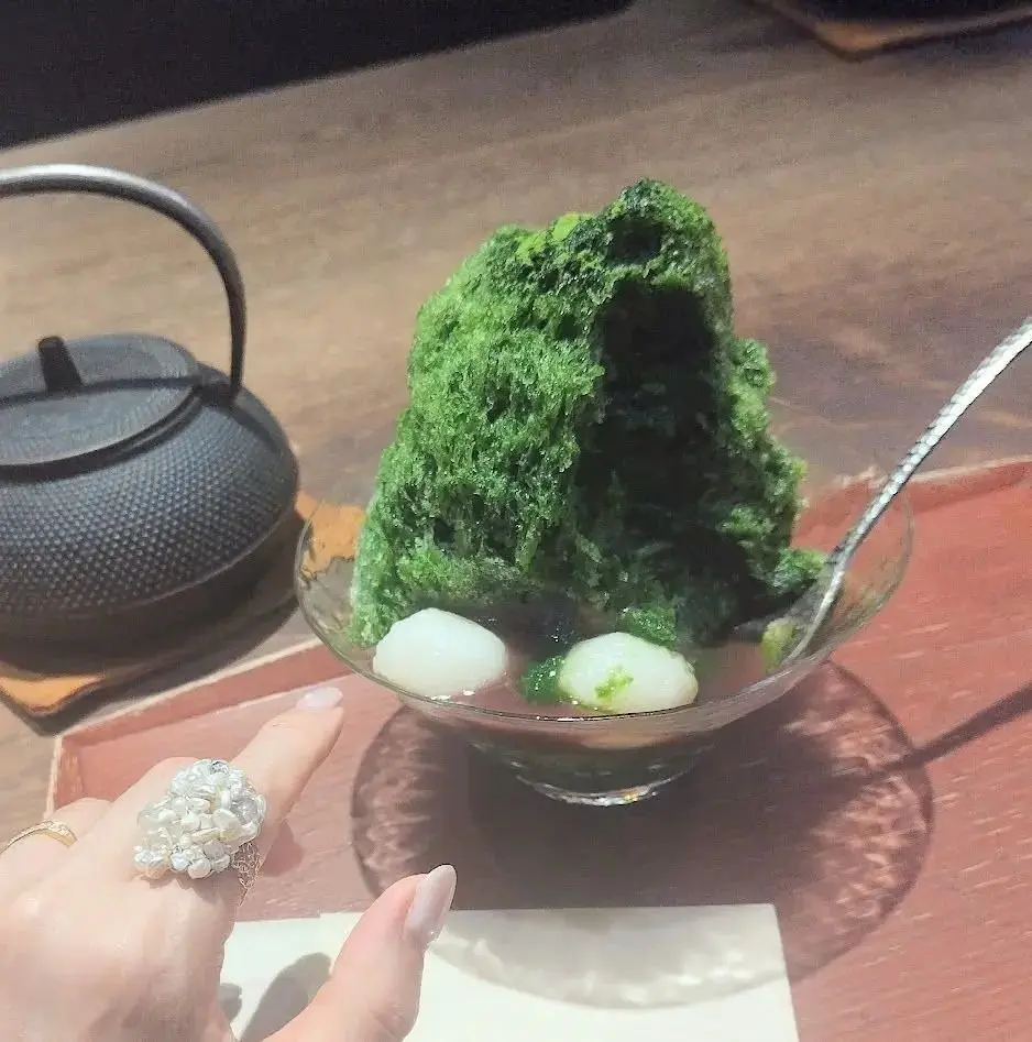 透明なガラスボウルに入った、緑色の苔のような盛り付けられた料理。中央には白い丸い団子風のものが2つ、緑色の料理の下に浮かんでいる。背景には鉄製の急須が置かれている。