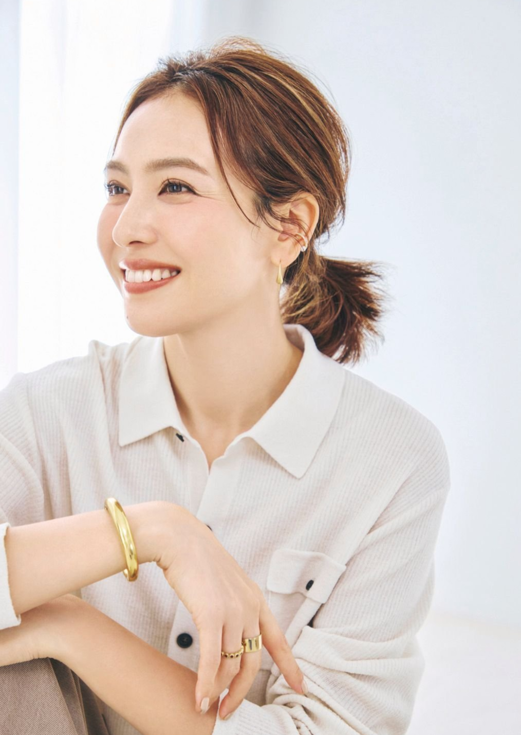 笑顔の女性が、白いシャツを着て座っているポートレート。茶色の肩長ヘアを後ろで束ね、ゴールドのバングルとリングを身に着けている。背景は白く、明るい自然光が差し込んでいる。