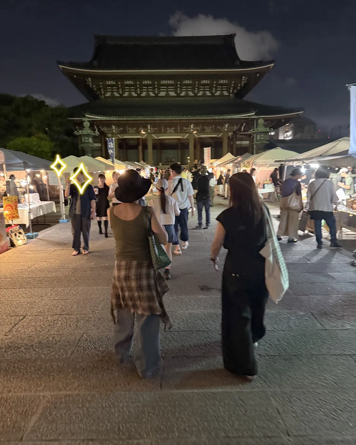 夜の市場で人々が歩いている様子。背景には、緑色の照明で照らされた伝統的な韓国建築の建物がそびえ立つ。手前には露店が並び、多くの人が散策している。