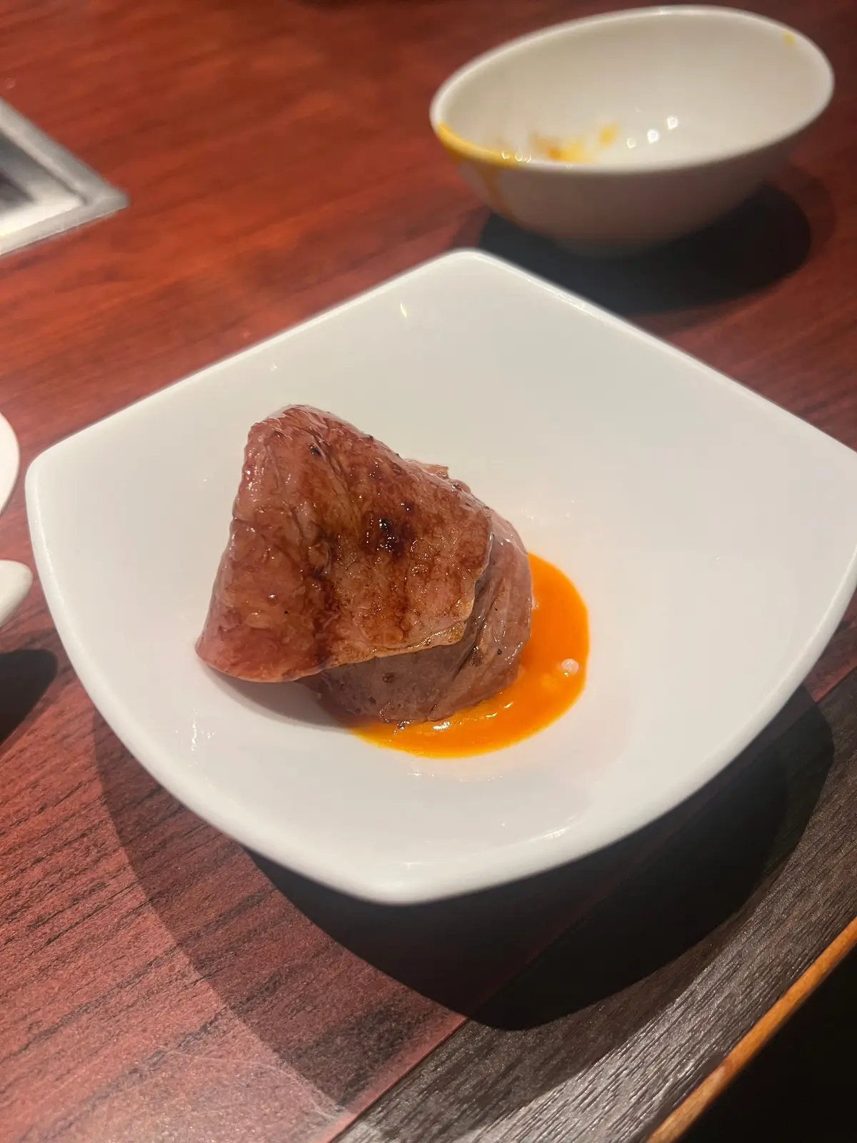 白磁の正方形皿に盛り付けられた、赤茶色の肉塊とオレンジ色のソース。肉は不規則な形をしており、皿の中央に配置されている。