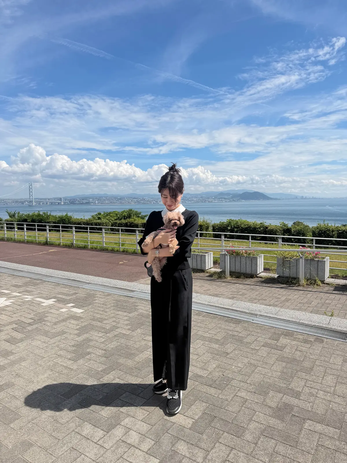 黒いワンピースを着た女性が、茶色の犬を抱いて立っている。背景には海と橋が見える、晴れた日の風景。