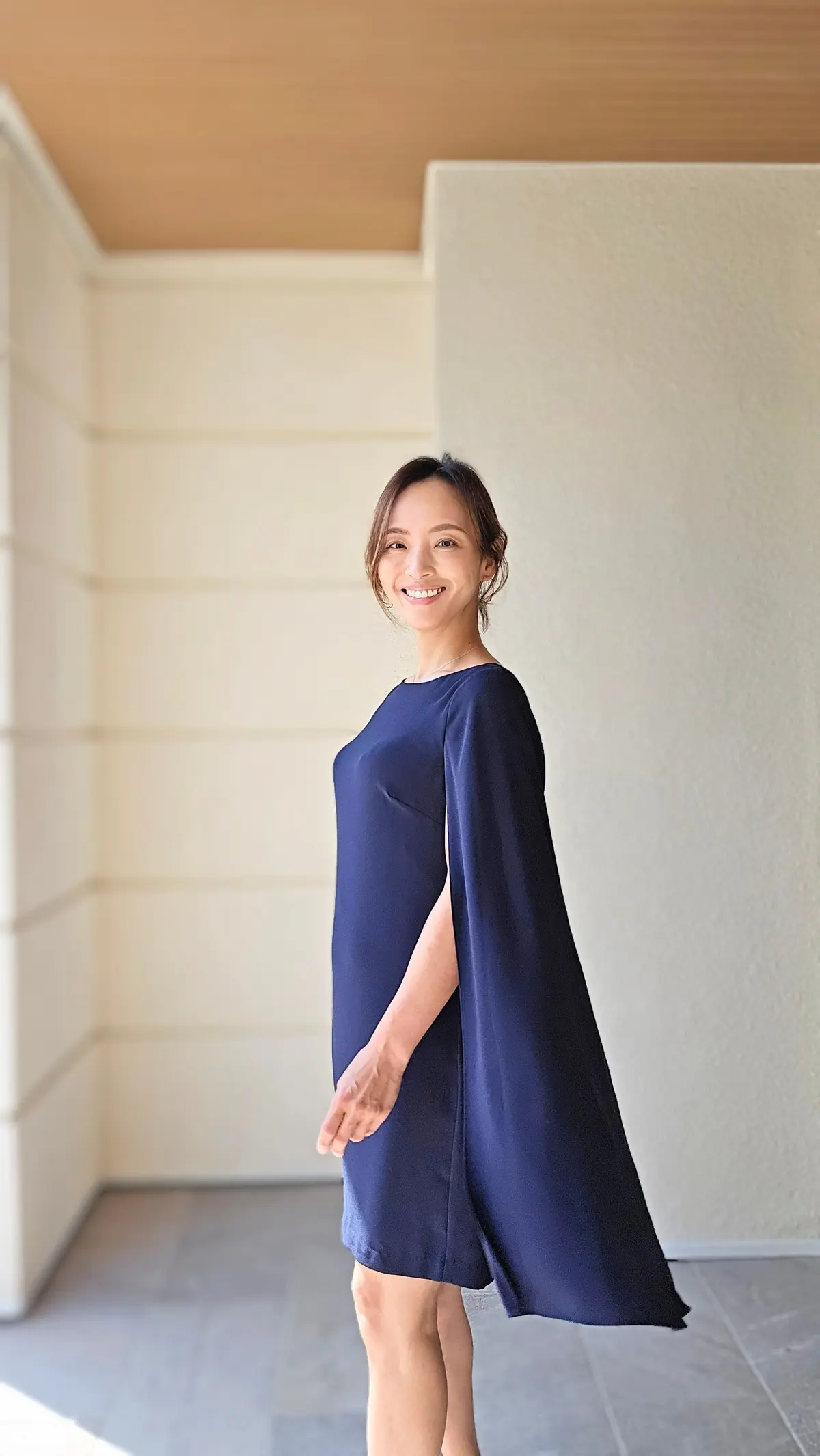 紺色のワンピースを着た女性が、笑顔でこちらを見ている。ワンピースは肩から流れるようなケープ付きで、膝丈のシンプルなデザイン。背景は白とベージュの壁で、明るい日差しが差し込んでいる。