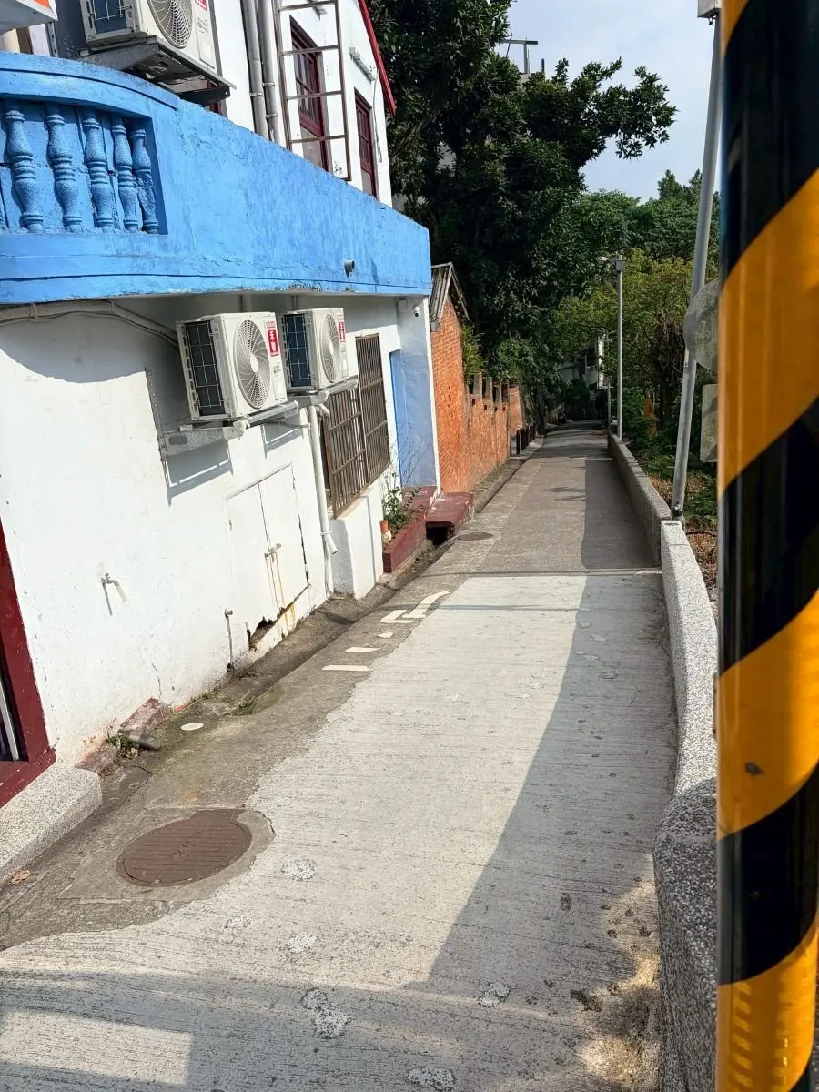 青いバルコニーとエアコン付きの白い建物が並ぶ狭い路地で、両側に木々が生い茂る