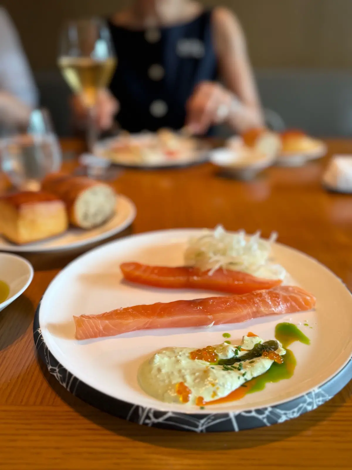 白黒の皿に盛り付けられた、サーモンとコールスロー、ソースの料理。背景にはワイングラスとパン、食事を楽しむ人の姿。