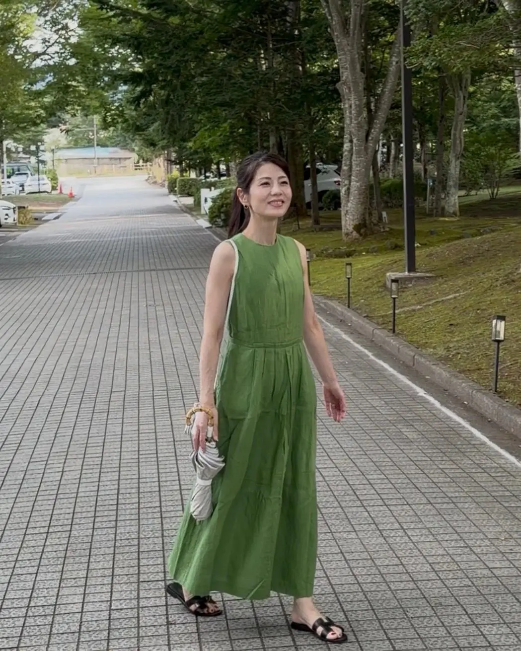 緑色のワンピースを着た女性が歩いている。黒いサンダルを履き、白い傘を持っている。背景には駐車場と道路が見える。