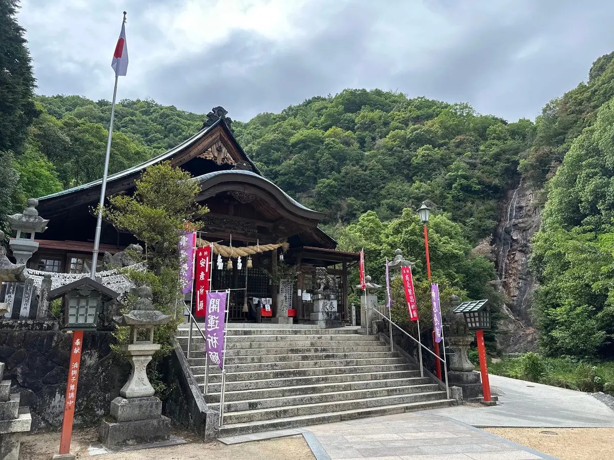 石段を登った場所に立つ伝統的な神社、緑豊かな山々が背景に、日本国旗が掲げられている
