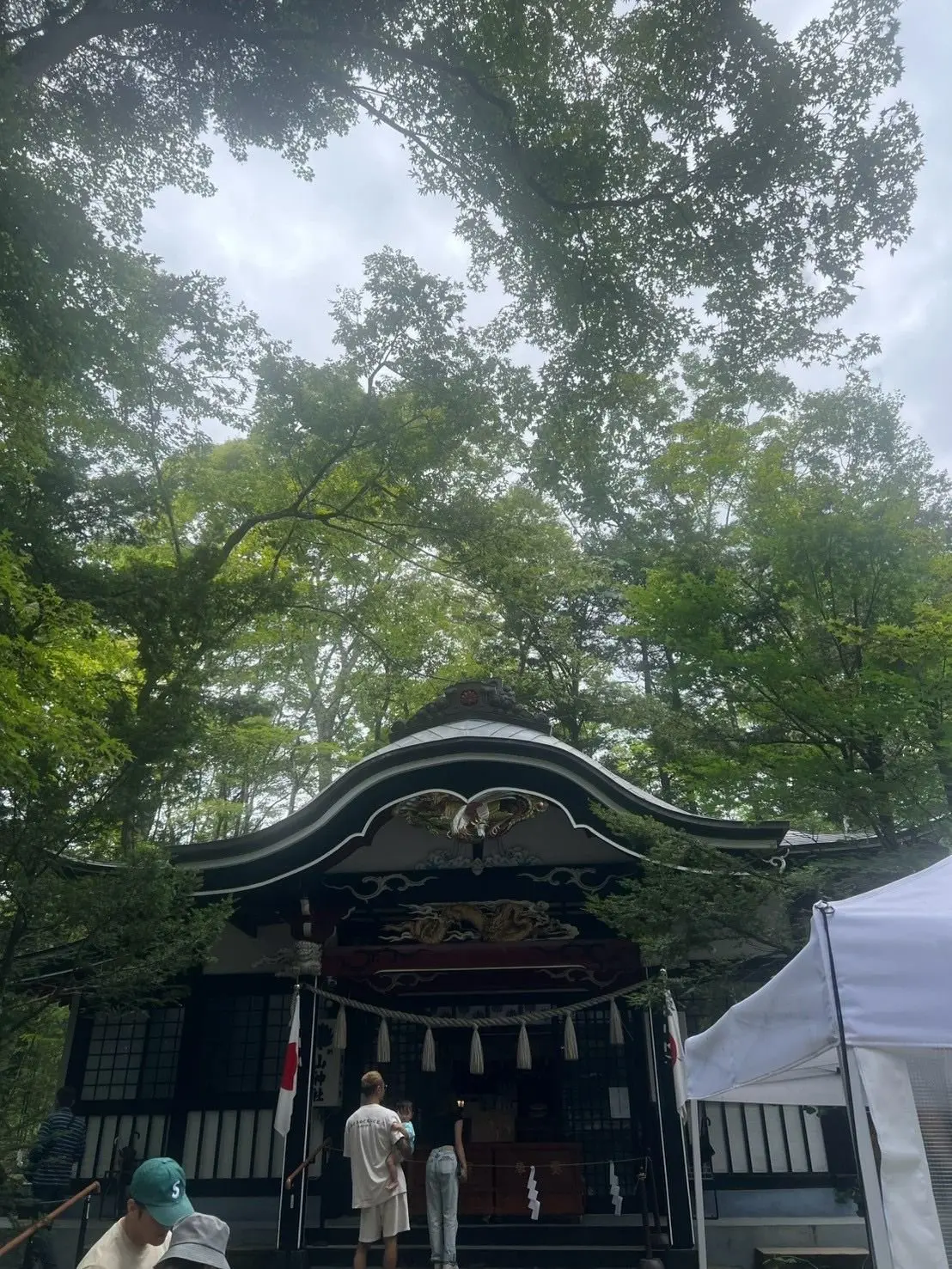 緑豊かな木々に囲まれた、黒い屋根の神社の建物。中央には赤い装飾があり、鳥の彫刻が施されている。手前には白いテントと人々が立っている。