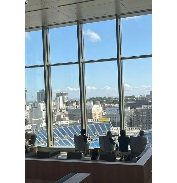 高層階の窓から見える、晴れた日の都市の風景。窓辺には座席が並び、人々がくつろいでいる。窓の外には青い空と白い雲、そして遠くに建物や緑が見える。スタジアムの座席の一部も見える。